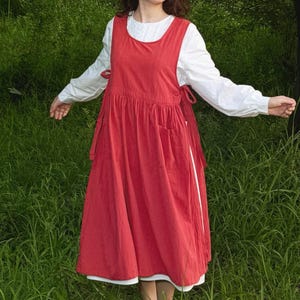 Puede incluir: Un vestido con delantal rojo con una camiseta interior blanca. El vestido tiene cuello redondo, bolsillos y lazos laterales. El vestido llega a la rodilla y está hecho de tela suave. Las mangas de la camiseta interior son largas y blancas.