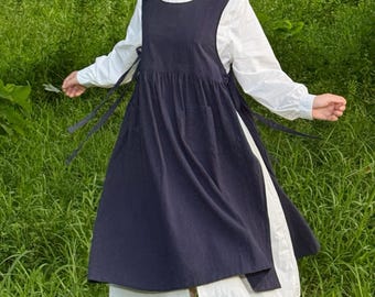 May include: A navy blue apron dress with a white long-sleeved blouse underneath. The apron dress has a scoop neckline and ties at the sides. The model is standing in a field of green grass.