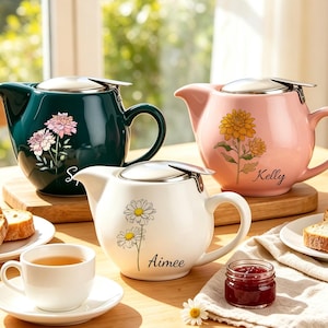 May include: Three ceramic teapots in dark green, light pink, and white, each featuring a floral illustration and a name: Sarah, Kelly, and Aimee. Each teapot has a silver-toned metal lid and handle. A white teacup and saucer are also present.