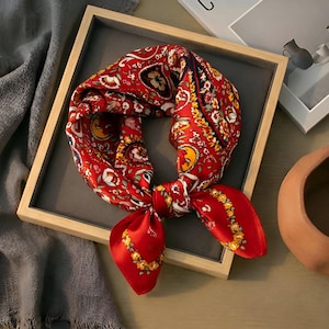 May include: A red silk scarf with a detailed floral and paisley pattern. The scarf is knotted and displayed in a wooden frame. The design includes yellow, white, and blue accents.