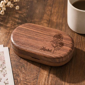 May include: An oval-shaped wooden box with a dark brown grain pattern. The lid features a laser-engraved illustration of a flower and the name Rachel in cursive script. The box has a smooth, polished finish and a horizontal seam along the side.