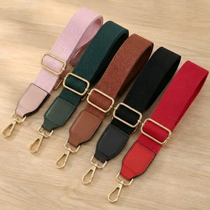 May include: Five colourful bag straps in pink, green, brown, black, and red. Each strap has a gold-coloured metal buckle and clasp, with leather or faux leather accents at the ends. The straps are arranged diagonally on a light wood surface.