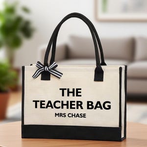 Peut inclure: Sac fourre-tout crème avec garniture et poignées noires. Le sac porte l'inscription "THE TEACHER BAG MRS CHASE" en noir. Un nœud rayé noir et blanc est attaché en haut.