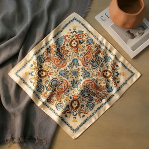 May include: Cream-coloured square scarf featuring a detailed paisley and floral pattern in orange, blue, and yellow. The scarf has a blue floral border. It is folded on a wooden surface next to a grey cloth.