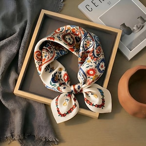 May include: A cream silk scarf featuring a colourful floral and paisley design in red, blue, and orange. The scarf is tied in a bow and presented in a wooden tray, highlighting its detailed pattern and sophisticated look.