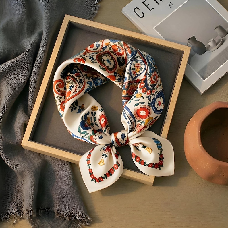 May include: A cream silk scarf featuring a colourful floral and paisley design in red, blue, and orange. The scarf is tied in a bow and presented in a wooden tray, highlighting its detailed pattern and sophisticated look.