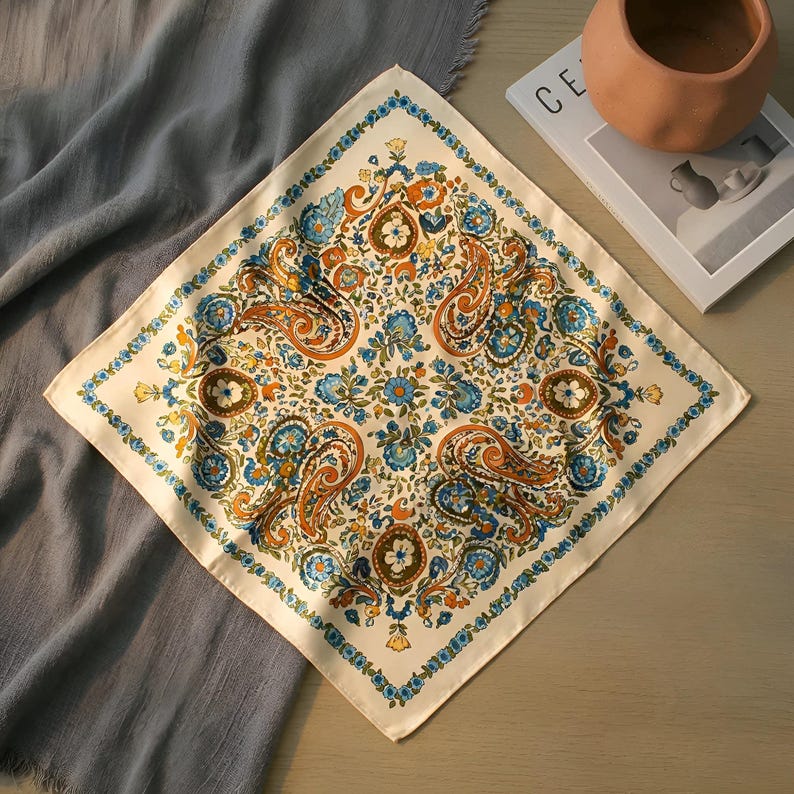 May include: Cream-coloured square scarf featuring a paisley and floral design in orange, blue, and green. The scarf has a detailed border and is folded on a grey surface. A terracotta pot and a magazine are in the background.