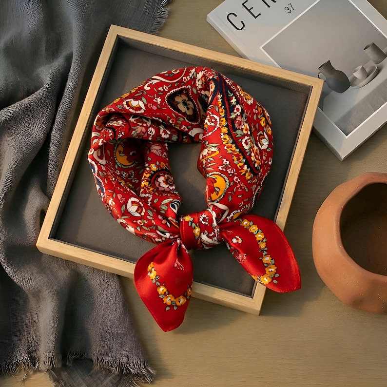May include: A red silk scarf with a detailed floral and paisley pattern. The scarf is knotted and displayed in a wooden frame. The design includes yellow, white, and blue accents.