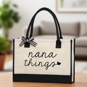 May include: Cream tote bag with black trim, handles, and the words "nana things" in a cursive font, with a heart. A black and white striped bow is attached to the top of the bag.