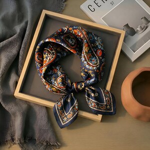 May include: A navy blue silk scarf with a paisley pattern in orange, red, and blue. The scarf is knotted and displayed in a wooden tray. The scarf is a square shape and has a decorative design.