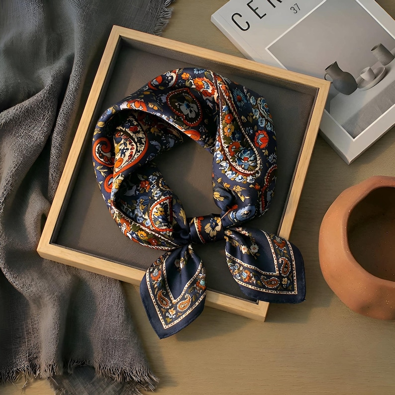 May include: A navy blue silk scarf with a paisley pattern in orange, red, and blue. The scarf is knotted and displayed in a wooden tray. The scarf is a square shape and has a decorative design.