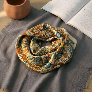 May include: A cream silk scarf with a paisley design in blue, orange, and yellow. The scarf is displayed on grey fabric, with a terracotta pot and an open book in the background. The scarf's pattern includes floral and swirl elements.