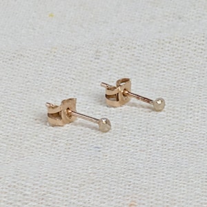 Op de afbeelding: Een paar kleine, goudkleurige oorstekers. Elke oorbel heeft een pin met een kleine, ronde, getextureerde knop. De oorbellen worden gepresenteerd op een wit, getextureerd oppervlak.