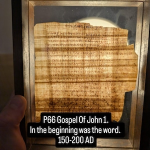 Könnte beinhalten: Gerahmtes Fragment des P66-Evangeliums nach Johannes 1, datiert auf 150-200 n. Chr. Der antike Text, geschrieben auf gealtertem Papyrus, ist durch das Glas sichtbar. Der Text lautet: „Am Anfang war das Wort.“