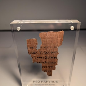 May include: A fragment of ancient papyrus, displayed in a clear acrylic case. The papyrus is a light brown color with visible text in black ink. The text is from the Gospel of John, dating from 125-150 AD. The case is secured with silver-colored fasteners.