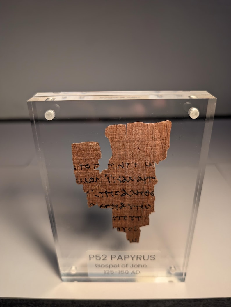 May include: A fragment of ancient papyrus, displayed in a clear acrylic case. The papyrus is a light brown color with visible text in black ink. The text is from the Gospel of John, dating from 125-150 AD. The case is secured with silver-colored fasteners.