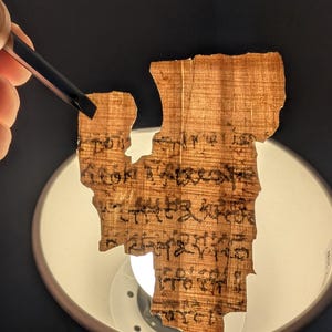 May include: A fragment of ancient papyrus with faded black ink script. The papyrus is held by tweezers, revealing its aged, textured surface. The background is dark, with a bright light illuminating the papyrus.