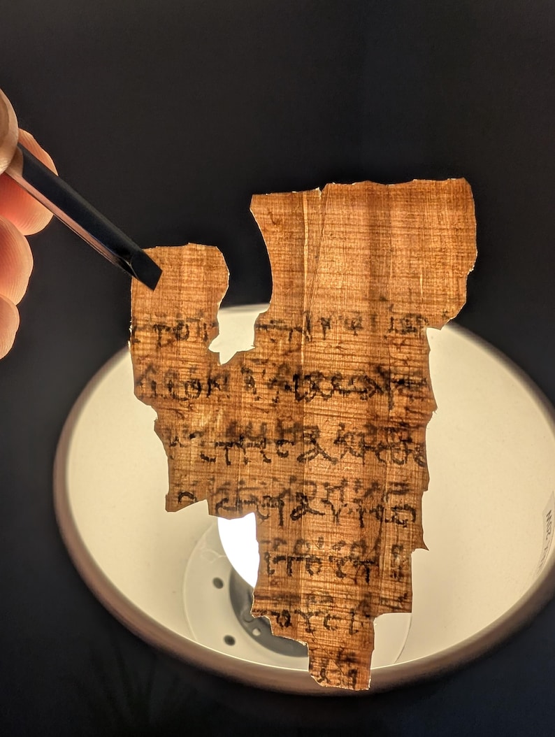 May include: A fragment of ancient papyrus with faded black ink script. The papyrus is held by tweezers, revealing its aged, textured surface. The background is dark, with a bright light illuminating the papyrus.