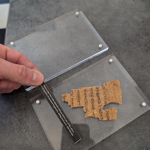 May include: A fragment of ancient text, likely a manuscript, displayed within a clear acrylic case. The text is in a faded brown color, with visible lettering. A small card with text is held near the fragment. The text on the card reads: Manuscript P52 (John Rylands Library 457) is one of the oldest surviving New Testament fragments, dating back to c. 125-150 AD.