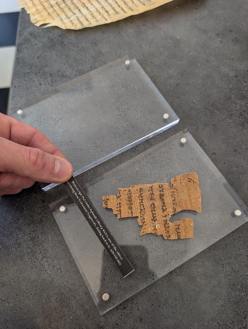 May include: A fragment of ancient text, likely a manuscript, displayed within a clear acrylic case. The text is in a faded brown color, with visible lettering. A small card with text is held near the fragment. The text on the card reads: Manuscript P52 (John Rylands Library 457) is one of the oldest surviving New Testament fragments, dating back to c. 125-150 AD.