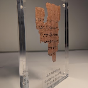 May include: A fragment of ancient papyrus with Greek text, encased in a clear acrylic display. The papyrus is a light brown color, with visible text in black ink. The display also includes text indicating it is P52 Papyrus, Gospel of John 18, from 125-150 AD.