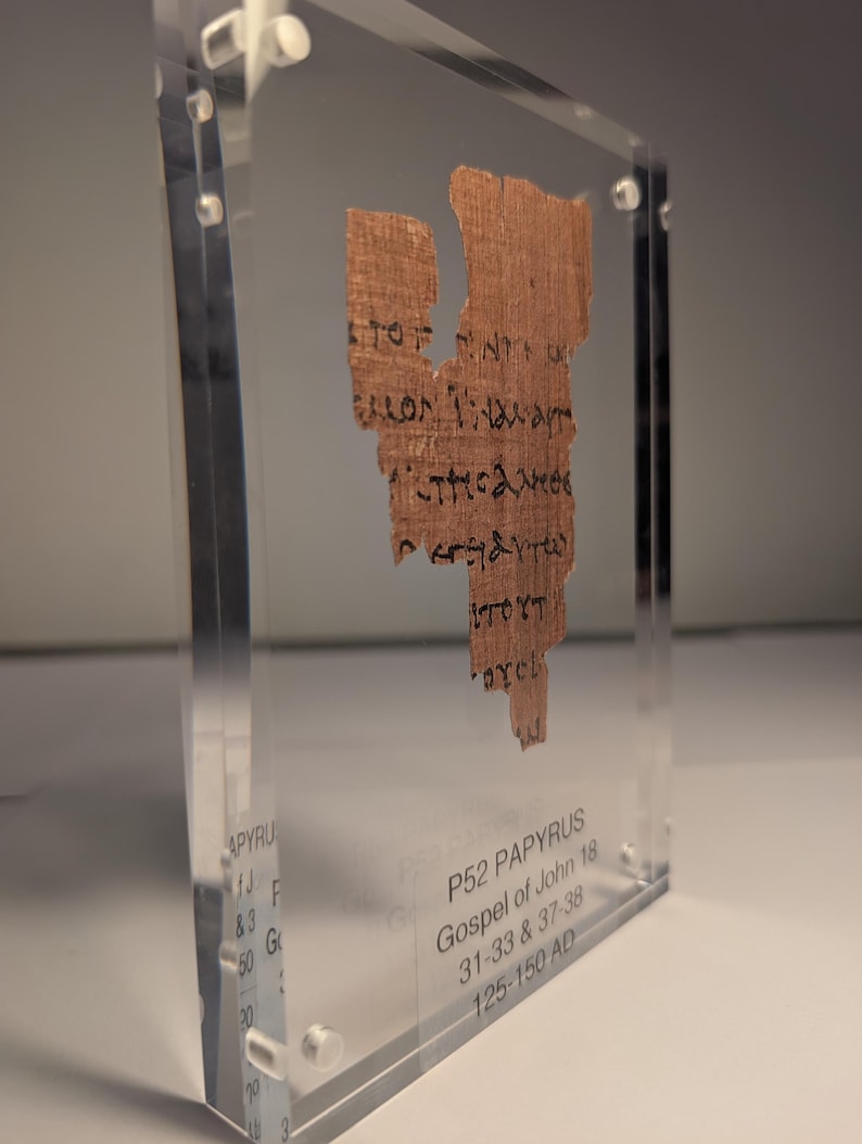 May include: A fragment of ancient papyrus with Greek text, encased in a clear acrylic display. The papyrus is a light brown color, with visible text in black ink. The display also includes text indicating it is P52 Papyrus, Gospel of John 18, from 125-150 AD.