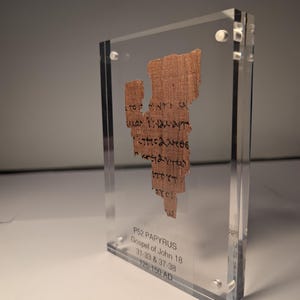May include: A fragment of ancient papyrus, P52, Gospel of John 18, displayed in a clear acrylic case. The papyrus is a light brown color with black handwritten text. The case has the text "P52 PAPYRUS Gospel of John 18 31:33 & 37-38 125-150 AD".