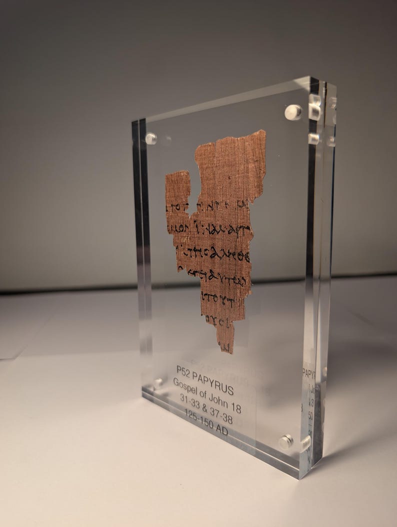 May include: A fragment of ancient papyrus, P52, Gospel of John 18, displayed in a clear acrylic case. The papyrus is a light brown color with black handwritten text. The case has the text "P52 PAPYRUS Gospel of John 18 31:33 & 37-38 125-150 AD".
