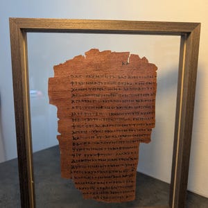 Könnte beinhalten: Ein gerahmtes Fragment aus altem Pergament mit verblasstem schwarzem Text. Das Pergament hat eine warme braune Farbe und wird in einem Holzrahmen mit einer transparenten Glasabdeckung präsentiert. Der Text ist in einer alten Sprache verfasst.
