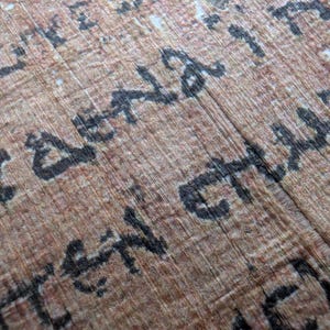 May include: Close-up of an ancient papyrus scroll with faded brown and tan tones. The papyrus has visible fibers and is inscribed with black, stylized text. The text appears to be in an ancient script, possibly hieroglyphs or a similar form of writing.