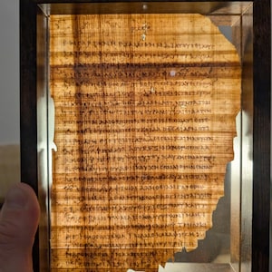 May include: A framed ancient papyrus fragment with visible text. The papyrus is a light brown color, with dark brown text. The frame is a dark brown wood, and the papyrus is lit from behind, creating a warm glow.