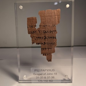 May include: A fragment of ancient papyrus, featuring text in black ink, is encased in a clear acrylic display. The papyrus is a light brown color and shows signs of age. The text on the papyrus is from the Gospel of John 18, dating from 125-150 AD.