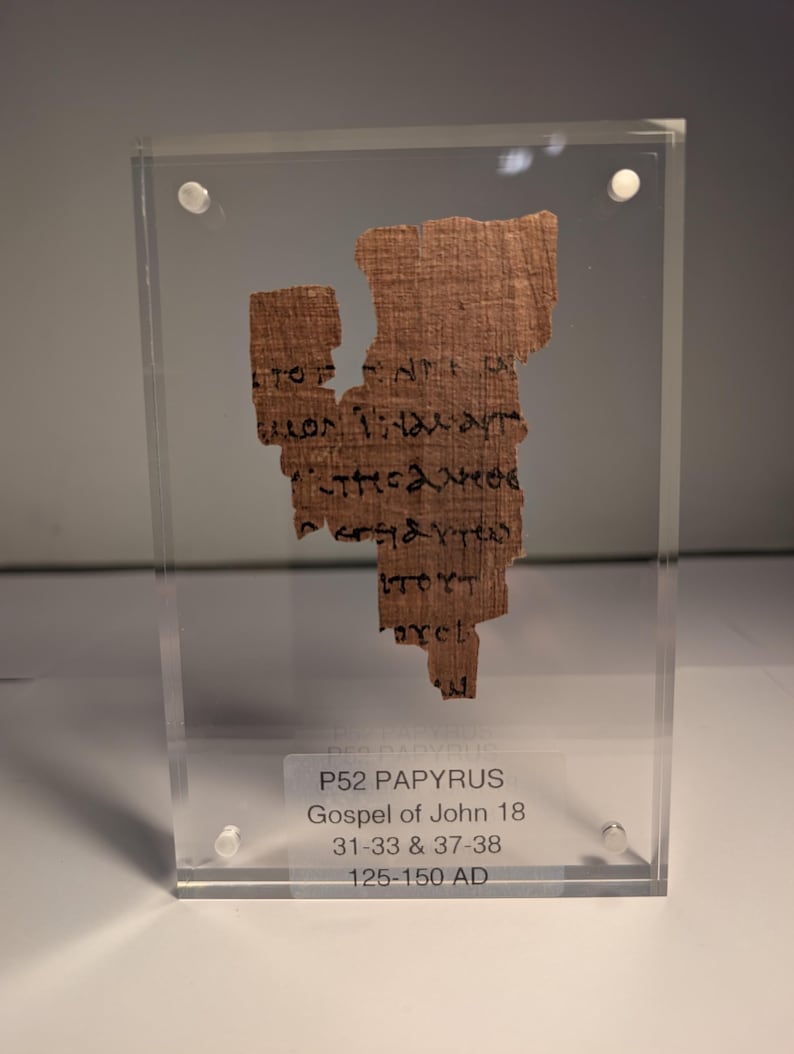 May include: A fragment of ancient papyrus, featuring text in black ink, is encased in a clear acrylic display. The papyrus is a light brown color and shows signs of age. The text on the papyrus is from the Gospel of John 18, dating from 125-150 AD.