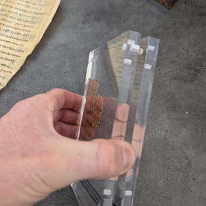 May include: A clear acrylic display case with a triangular shape, held by a hand. The case has multiple layers and small white fasteners. Inside the case are pieces of aged paper with visible text. The background includes a piece of aged paper with text.