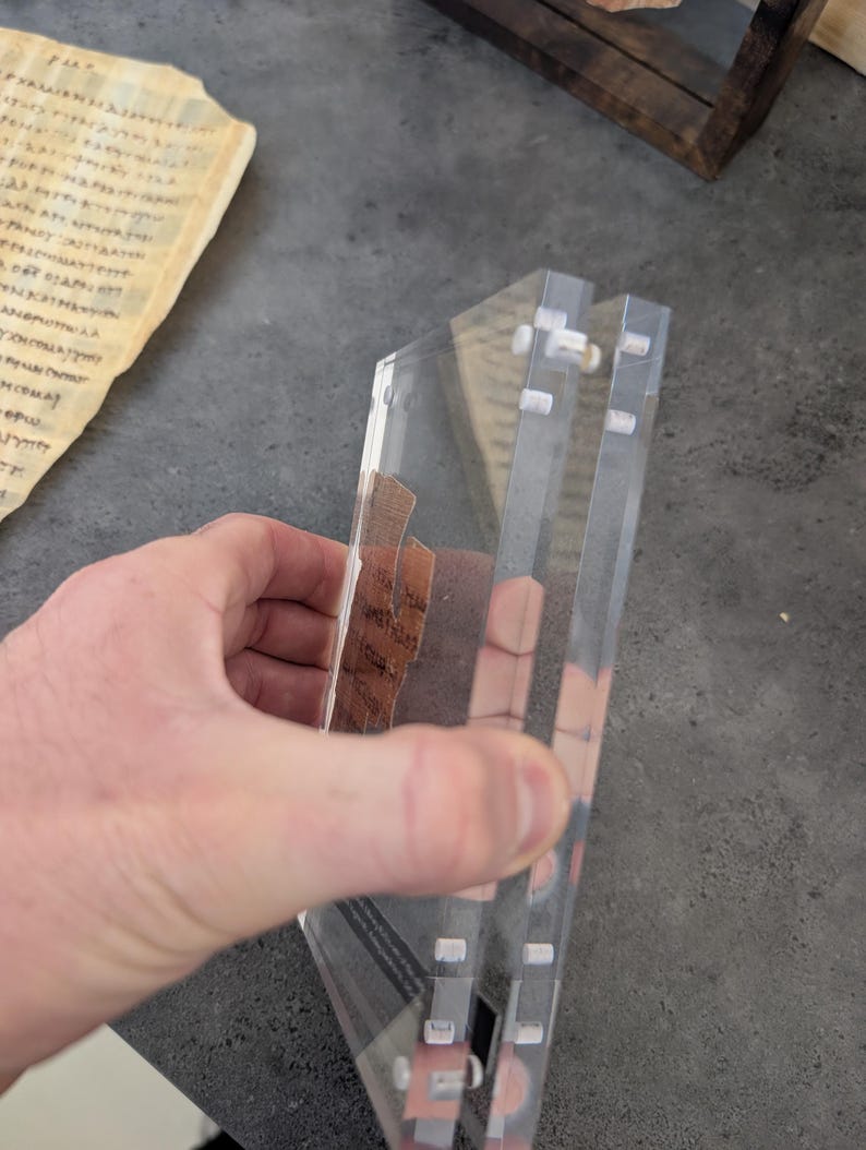 May include: A clear acrylic display case with a triangular shape, held by a hand. The case has multiple layers and small white fasteners. Inside the case are pieces of aged paper with visible text. The background includes a piece of aged paper with text.