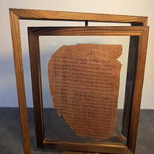 May include: A framed ancient papyrus fragment. The papyrus, with visible text, is displayed within a clear glass pane, encased by a wooden frame. The frame is a warm brown colour, and the papyrus has a tan hue.