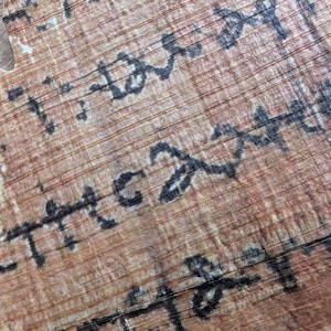 May include: Close-up of a fragment of ancient papyrus with faded black ink script. The papyrus has a light brown color with visible horizontal and vertical lines. The script appears to be in an ancient language.