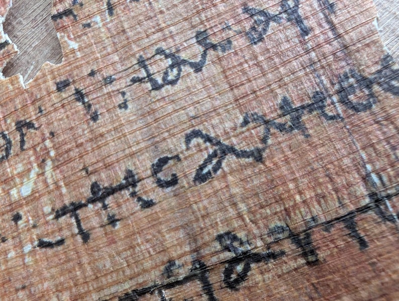 May include: Close-up of a fragment of ancient papyrus with faded black ink script. The papyrus has a light brown color with visible horizontal and vertical lines. The script appears to be in an ancient language.