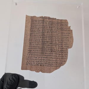 May include: A fragment of ancient papyrus featuring handwritten Greek text in dark ink, preserved between two clear acrylic sheets. The papyrus has a rough, uneven edge and a light brown, fibrous texture, displaying multiple lines of script.