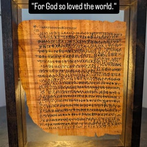 Può includere: Antico papiro incorniciato con testo, con la citazione "Dio infatti ha tanto amato il mondo." da Giovanni 3:16. Il papiro è esposto in una cornice di legno scuro, mostrando la pergamena invecchiata e la scrittura a mano.