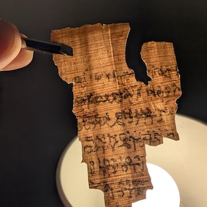 May include: A fragment of ancient papyrus with faded black text. The papyrus is held by tweezers, revealing its torn edges and aged appearance. The background is dark, with a bright light illuminating the papyrus.