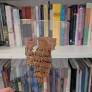 May include: A fragment of ancient manuscript P52, one of the oldest surviving New Testament fragments, dating back to c. 125-150 AD. The text is written in black ink on a tan papyrus. The manuscript is encased in a clear protective holder.
