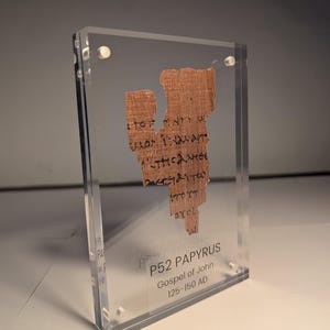 Könnte beinhalten: Ein Fragment des P52-Papyrus, Evangelium nach Johannes, aus der Zeit von 125-150 n. Chr., in einem transparenten Acrylglasgehäuse. Das Papyrus ist hellbraun mit sichtbarem Text in schwarzer Tinte. Das Acrylgehäuse wird von kleinen, silberfarbenen Schrauben zusammengehalten.