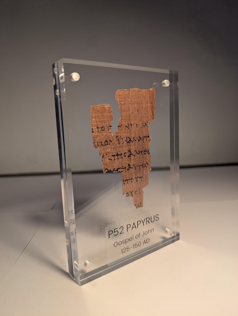 May include: A fragment of P52 Papyrus, Gospel of John, dating from 125-150 AD, encased in clear acrylic. The papyrus is a light brown color with visible text in black ink. The acrylic case is held together with small, silver-colored screws.