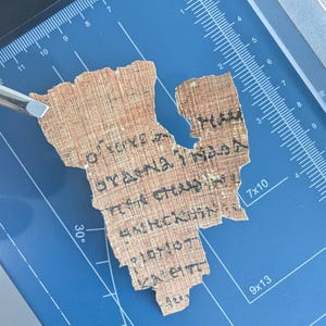 May include: A fragment of ancient papyrus with faded black text, possibly Greek or Coptic, is held by a silver tool on a blue cutting mat. The papyrus is a light brown color and appears to be fragile.