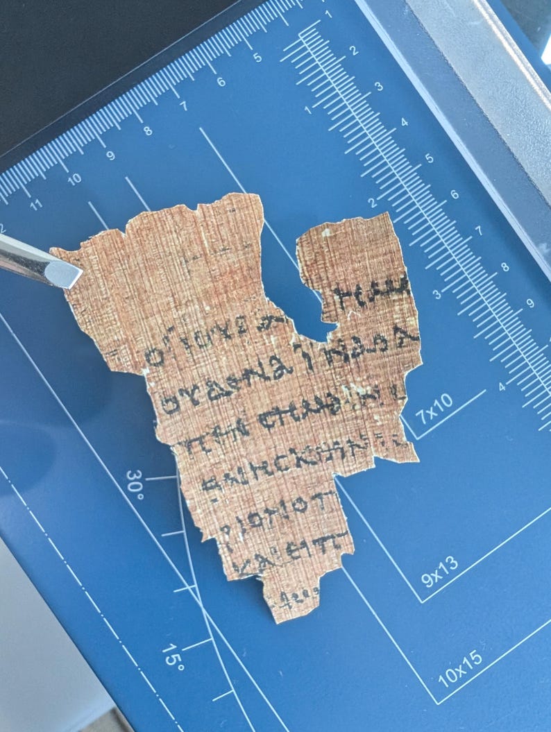 May include: A fragment of ancient papyrus with faded black text, possibly Greek or Coptic, is held by a silver tool on a blue cutting mat. The papyrus is a light brown color and appears to be fragile.