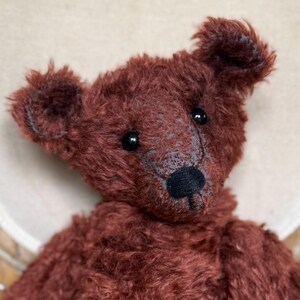 Puede incluir: Un oso de peluche hecho a mano, de un rico tono marrón rojizo. El oso tiene una nariz negra, ojos oscuros y una apariencia ligeramente desgastada. Está sentado en una silla de color crema, mostrando su suave y texturizada piel y almohadillas blancas.