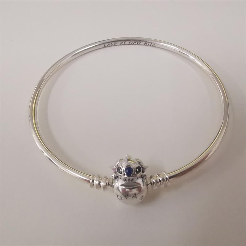 Puede incluir: Una pulsera de plata con un dije de personaje. El dije tiene un cuerpo plateado con una gema azul y otras gemas peque&ntilde;as. La pulsera tiene grabada la frase "Love at first bite".