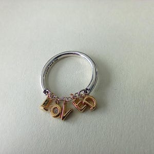 May include: A silver ring with gold-coloured charms spelling "LOVE" and a small diamond. The ring has a polished finish and is set against a neutral background. The charms are suspended from the ring.