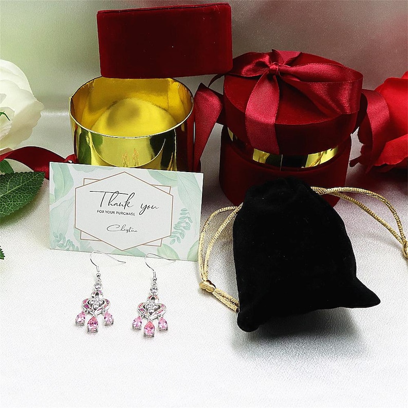 May include: A pair of silver earrings with pink teardrop-shaped stones. The earrings are displayed with a thank you card, a black velvet pouch, and red velvet jewelry boxes with gold accents.